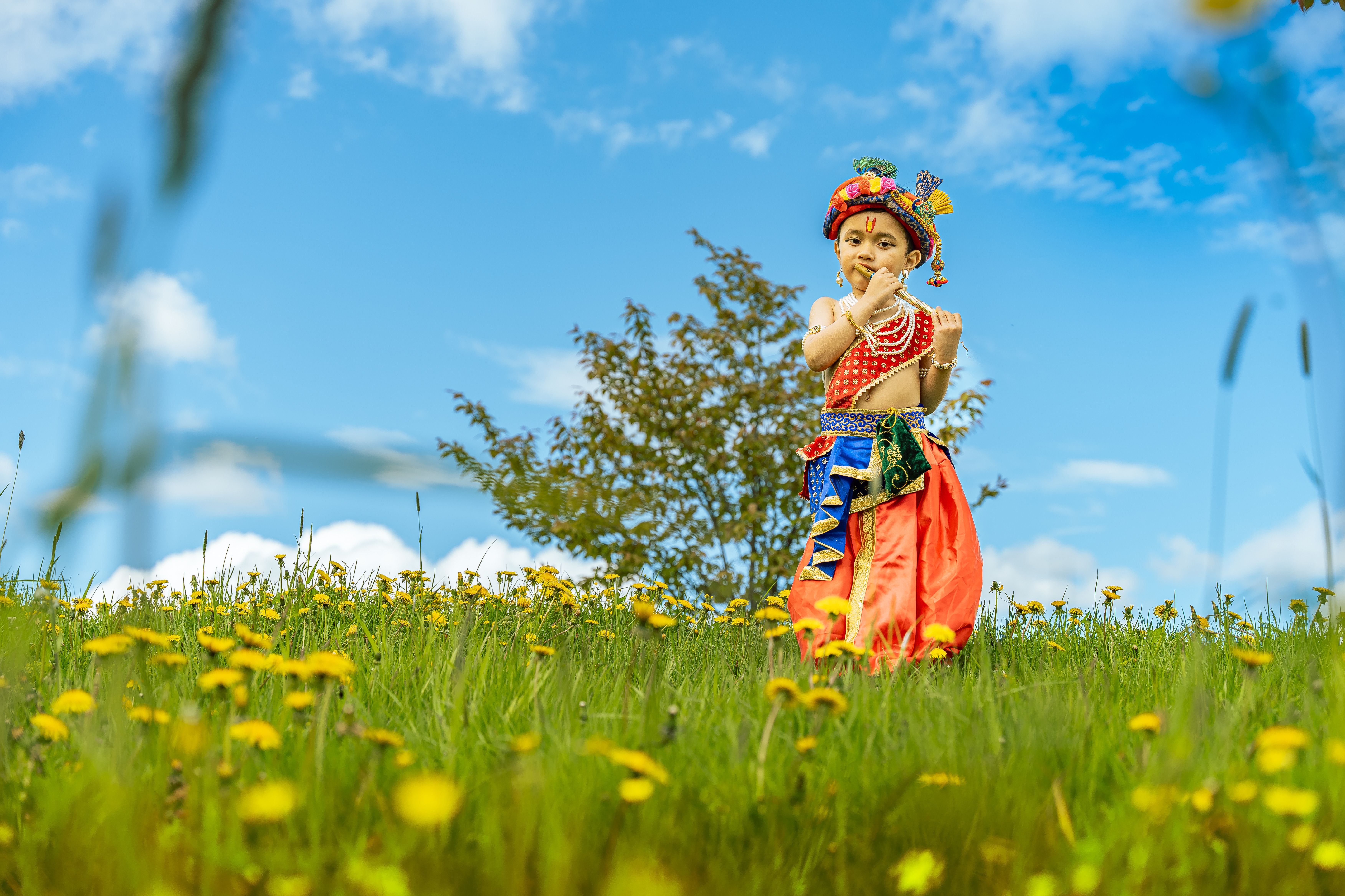 A child dressed as Lord Krishna in traditional attire poses in a scenic Swedish field under a clear blue sky.