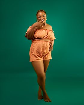 A confident woman poses in an orange summer outfit on a green background.