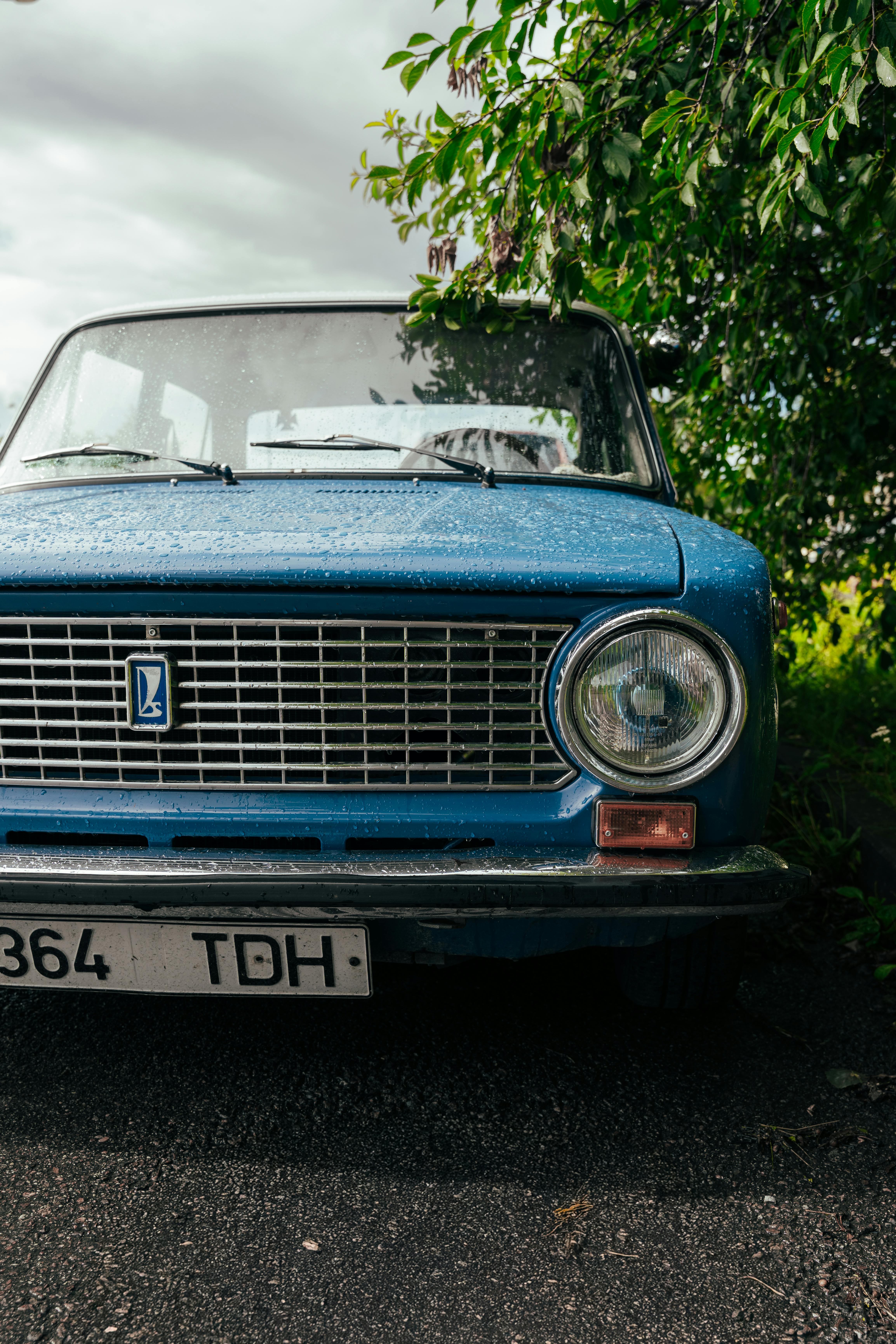 Close-up of a vintage blue Lada car parked outdoors under a tree with raindrops visible.