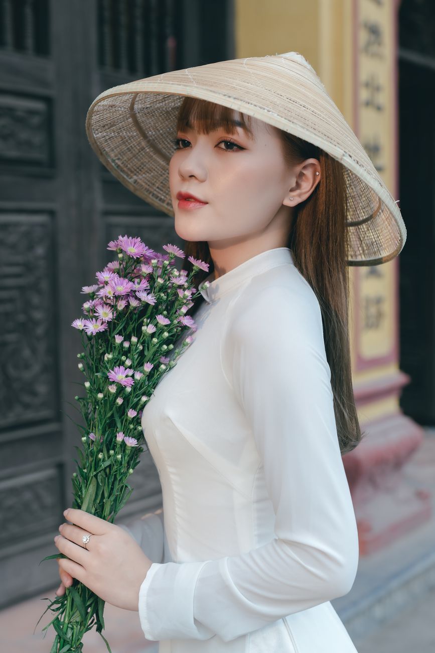 A young woman wearing a traditional Asian outfit and hat, holding a bouquet of flowers outdoors.