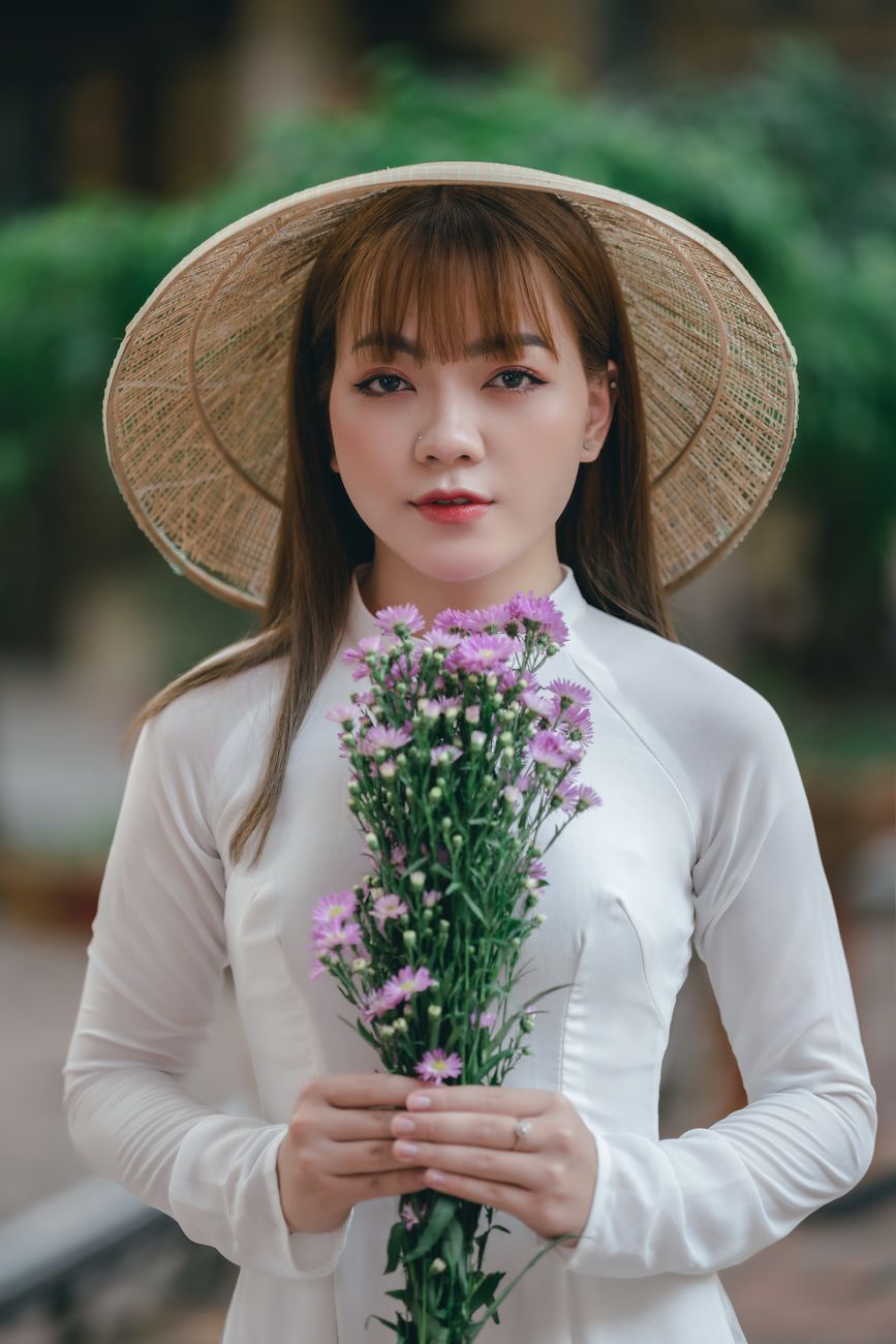 Young woman wearing a traditional straw hat and white dress holding pink flowers.