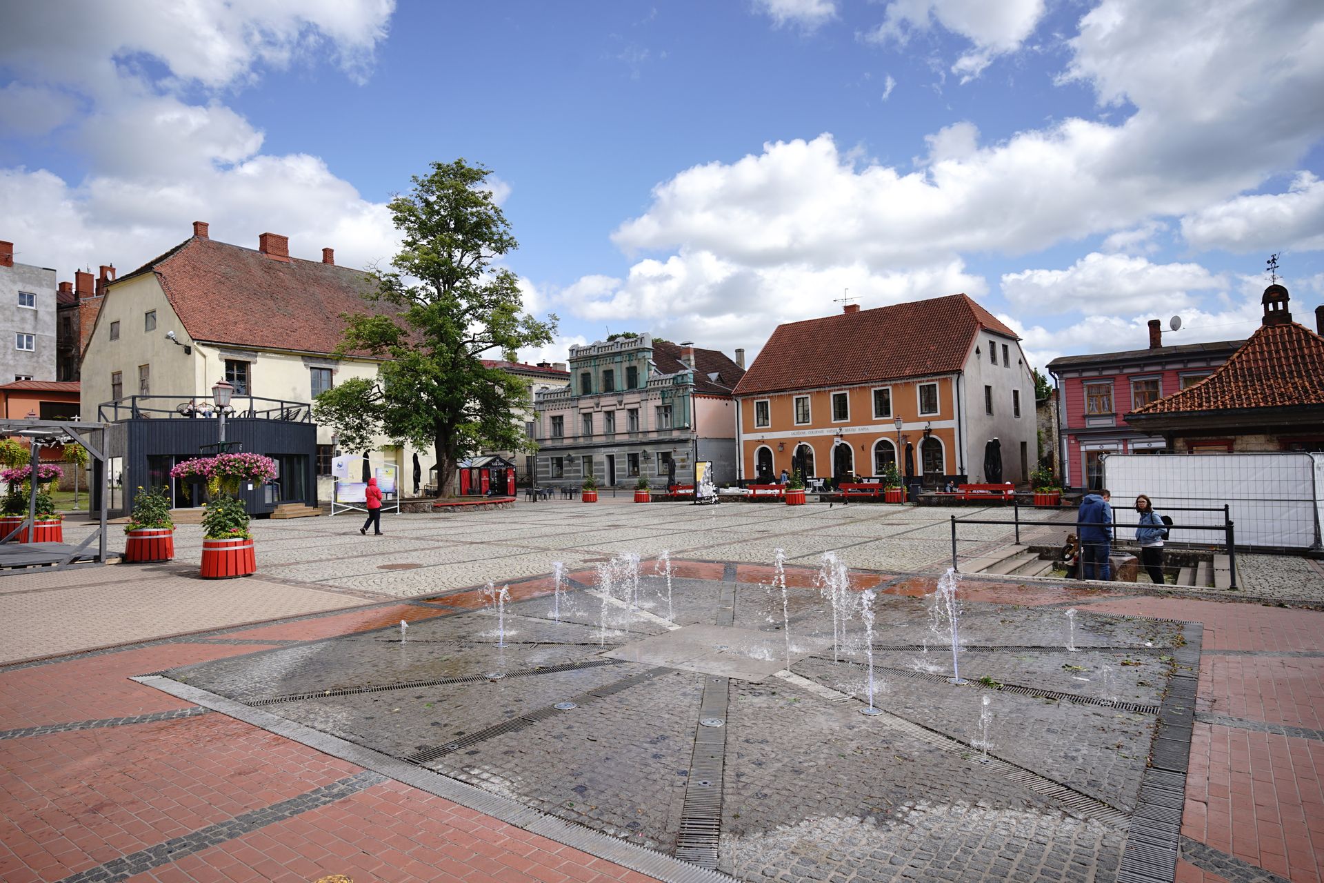 Paved square with a water fountain surrounded by historic European buildings and vibrant flowers.