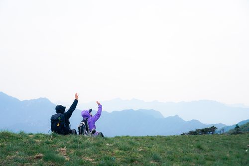Free Two hikers wave enthusiastically atop a grassy hill with mountain views. Stock Photo