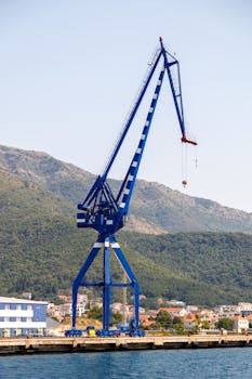 A towering blue crane stands at the waterfront, set against a mountainous landscape.