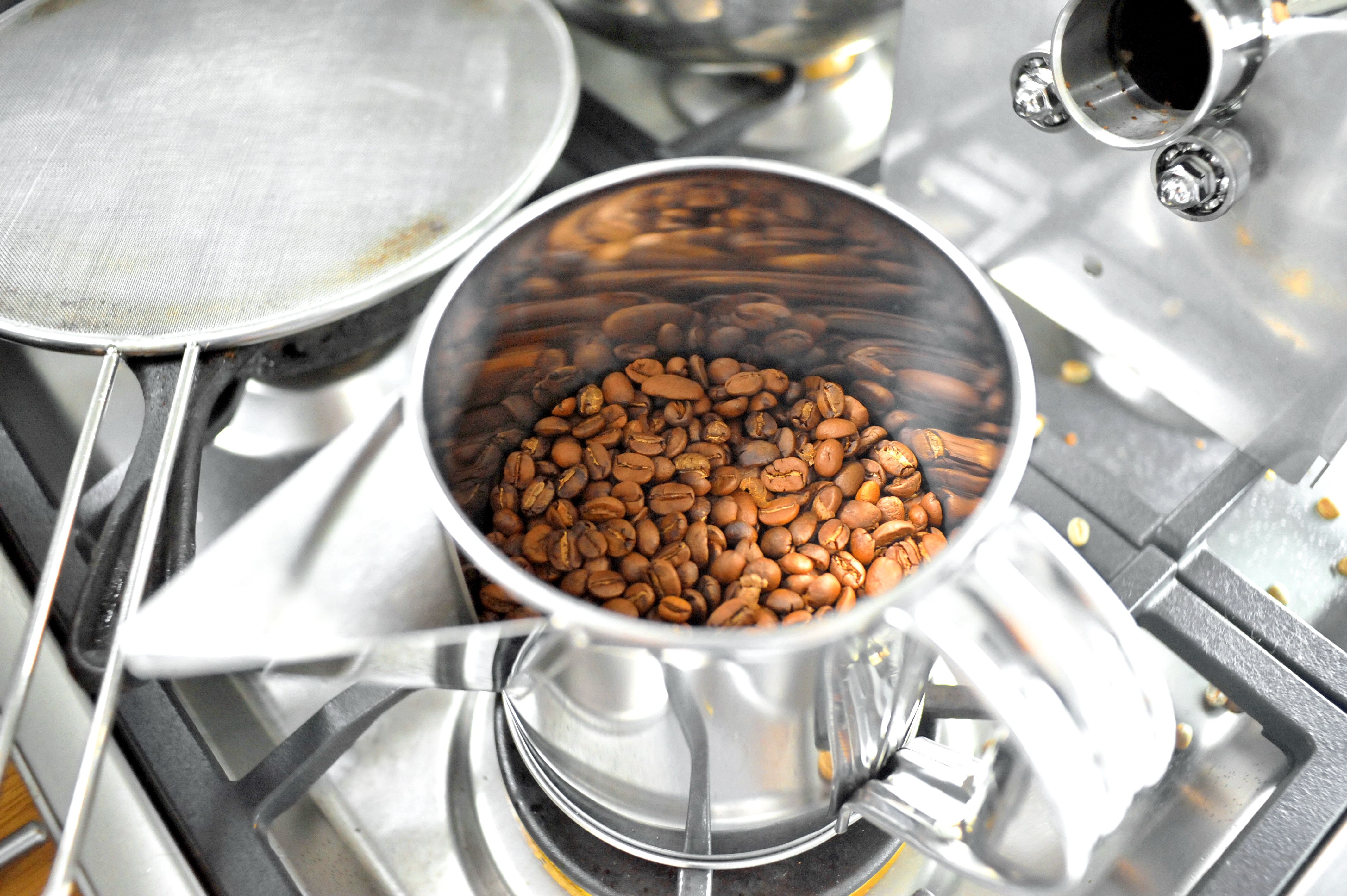 A stainless steel jug filled with freshly roasted coffee beans on a gas stove.