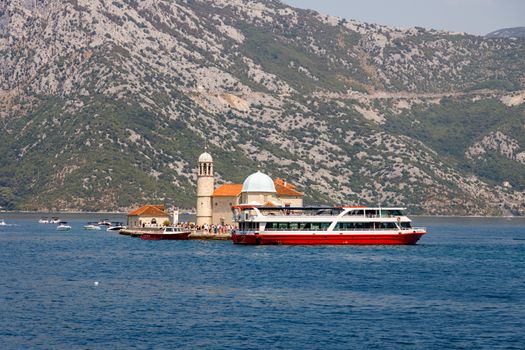 Tourist ferry near Our Lady of the Rocks in Montenegro with scenic mountain backdrop.