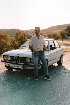 A man sits on a classic car in a sunlit landscape in İzmir, Türkiye.
