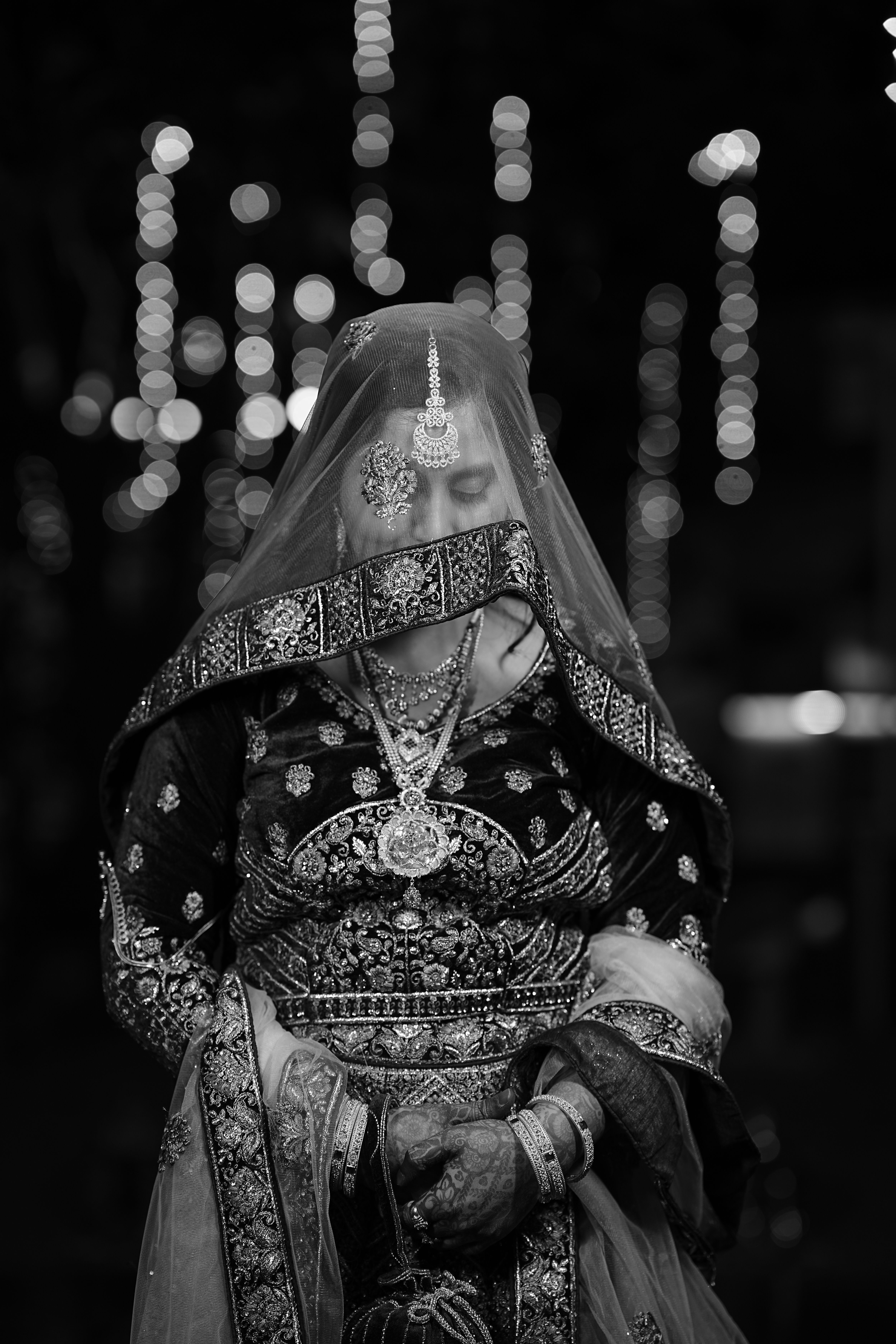 Elegant black and white portrait of a traditional bride in intricate attire with bokeh lights background.