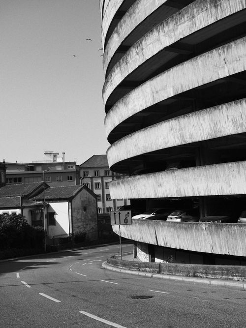 Free Black and white photo of a spiral parking garage beside old town buildings, capturing urban architecture. Stock Photo