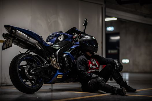 A motorcyclist in black gear sits beside a blue sportbike in a dimly lit garage.