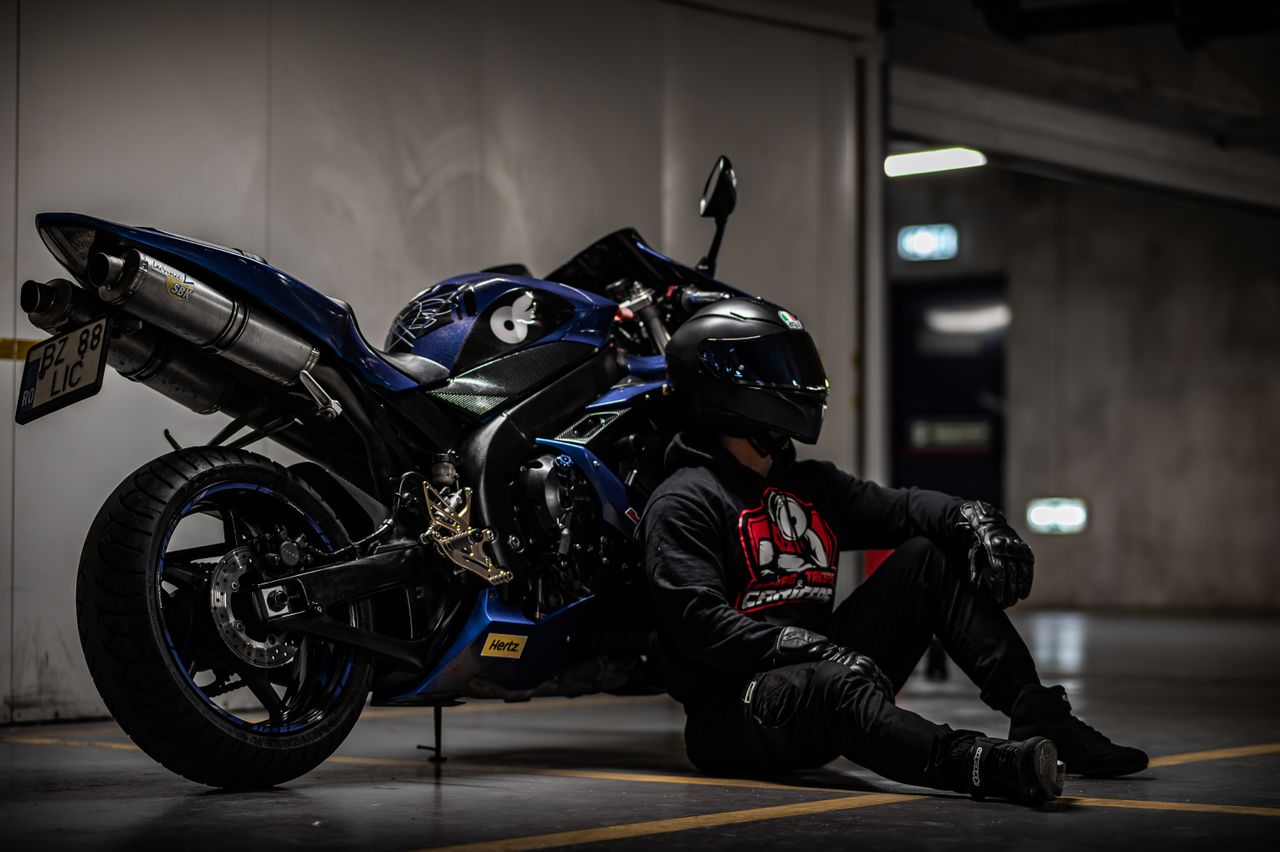 a motorcyclist in black gear sits beside a blue sportbike in a dimly l