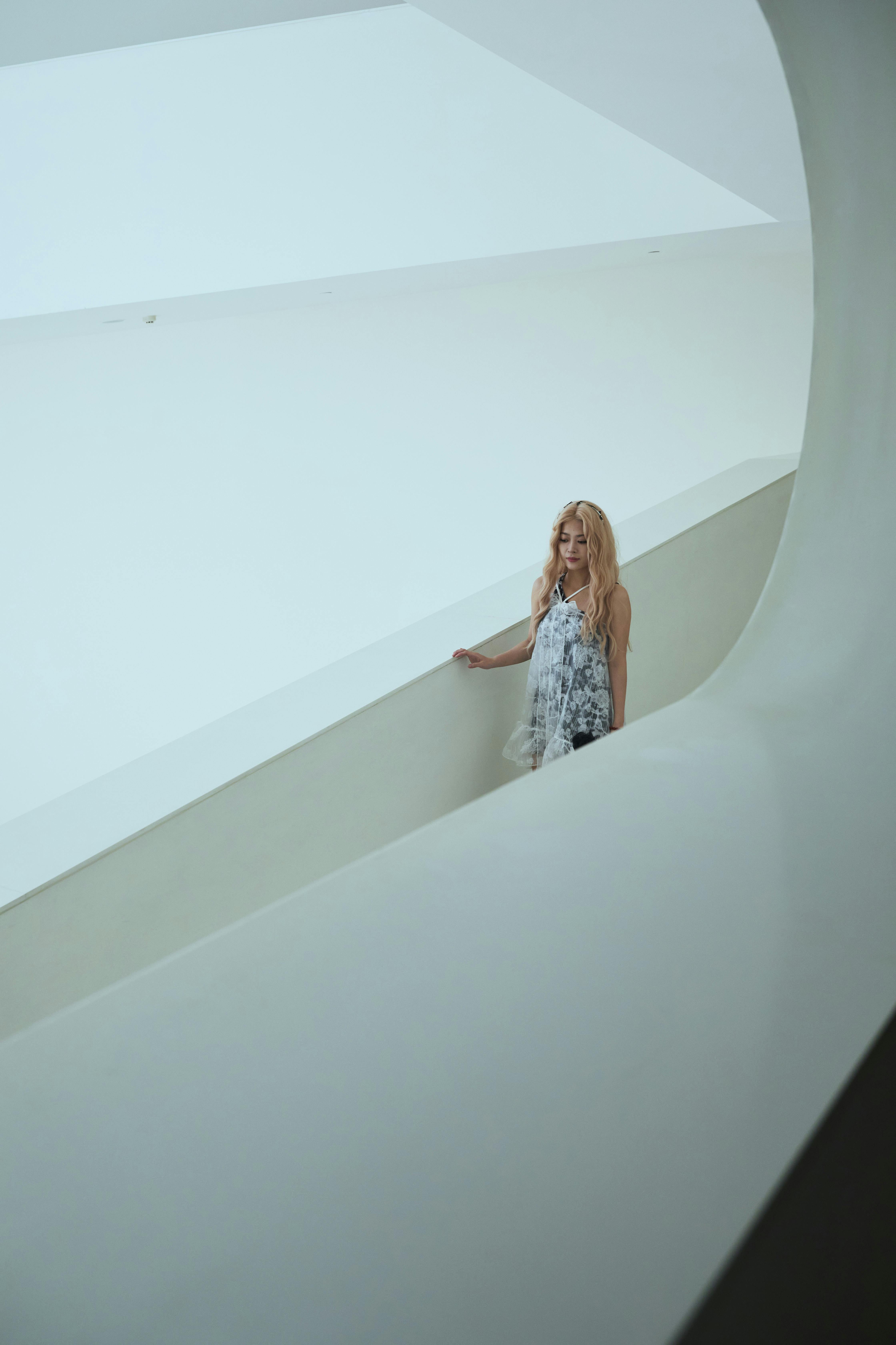 A thoughtful woman in flowing dress stands in a modern, minimalist architectural space.