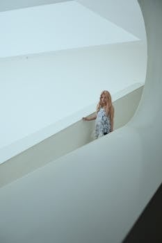 A thoughtful woman in flowing dress stands in a modern, minimalist architectural space.
