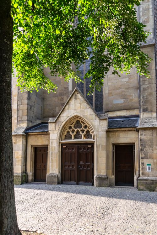 Free Gothic facade of a historic stone church with arched doorways and leafy tree shade. Stock Photo