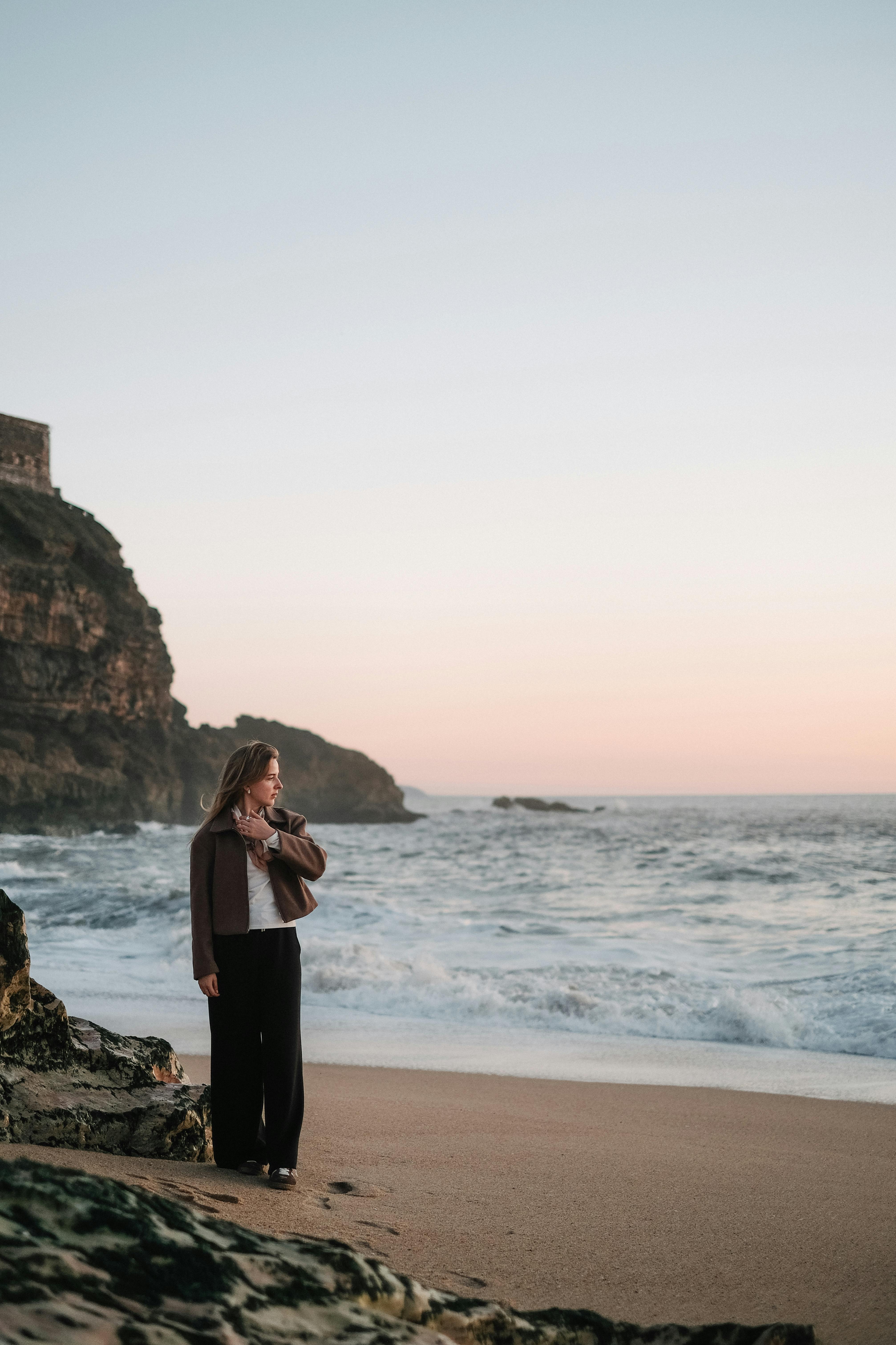 A woman stands on a sandy beach at sunset, near rocky cliffs and ocean waves.
