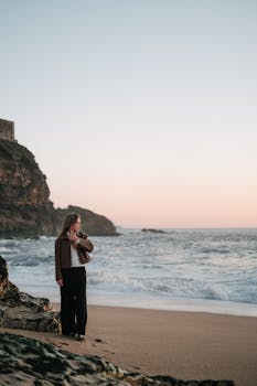 A woman stands on a sandy beach at sunset, near rocky cliffs and ocean waves.