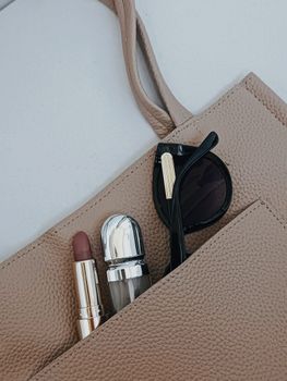 Elegant sunglasses, lipstick, and perfume in a beige handbag pocket.