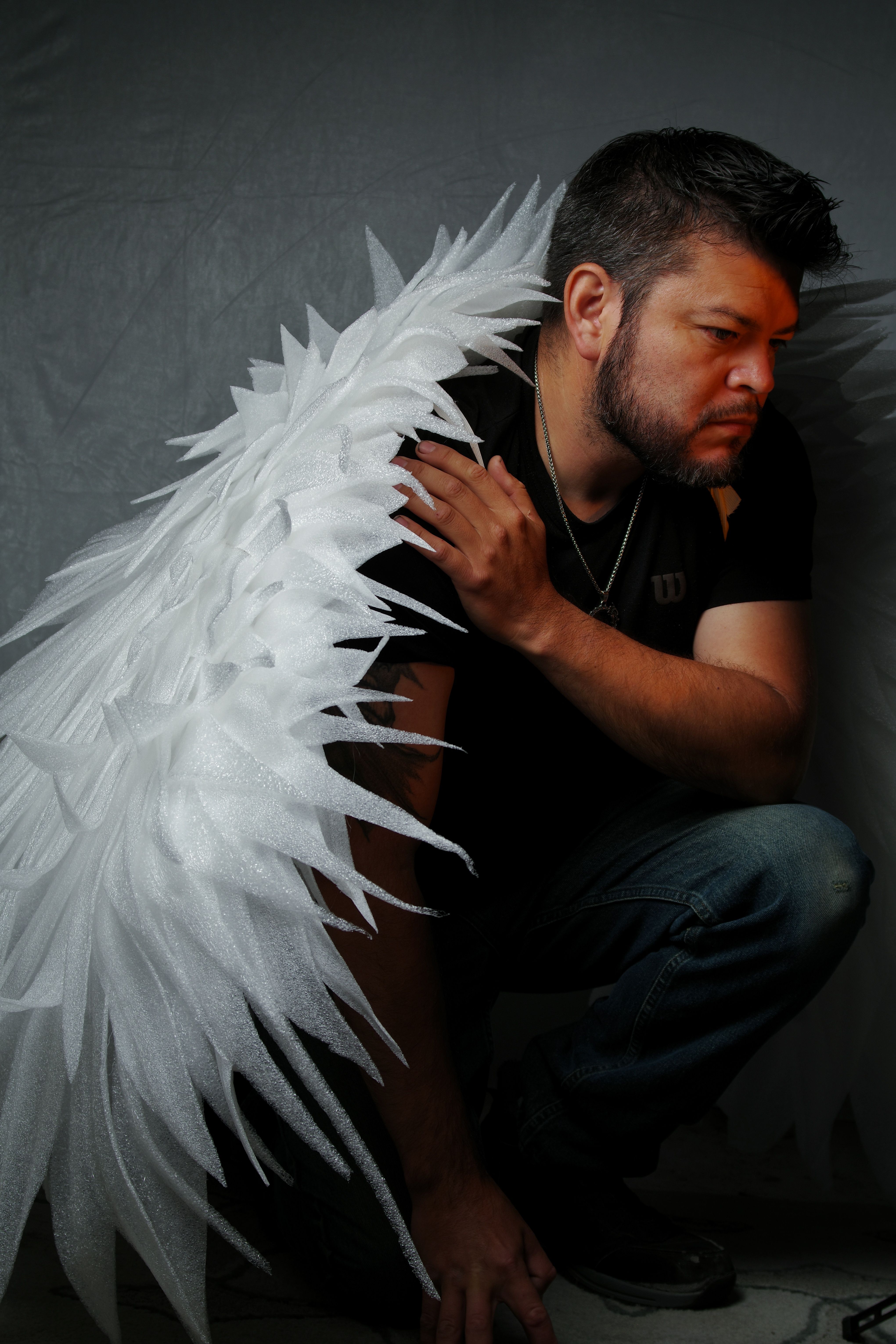 A man crouches dramatically with large angel wings in a moody setting.