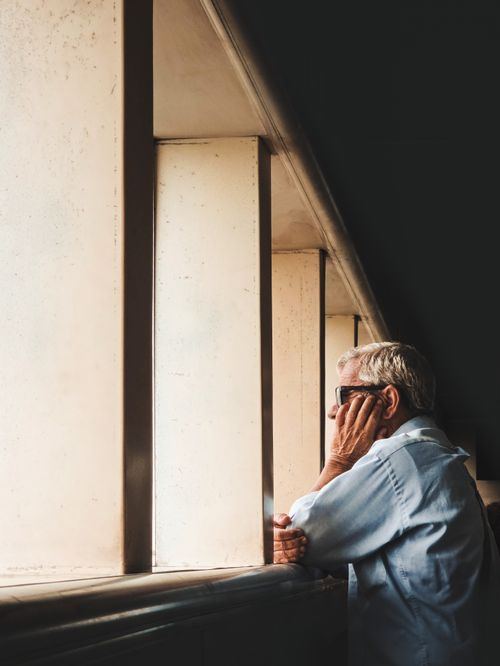 Free Senior man with glasses thoughtfully looking out of a large indoor window. Stock Photo