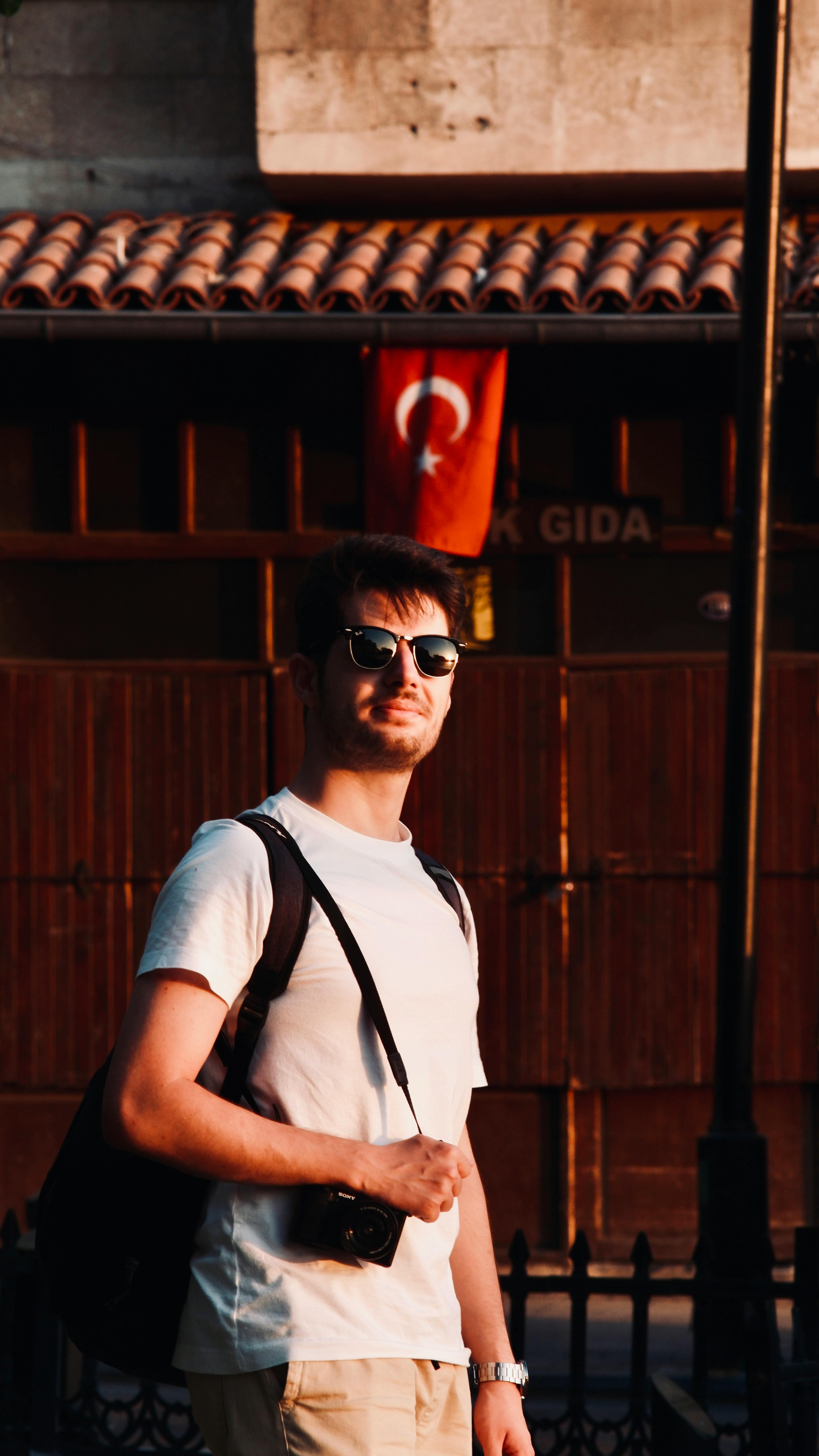 A man wearing sunglasses and a backpack poses near a Turkish flag during sunset.