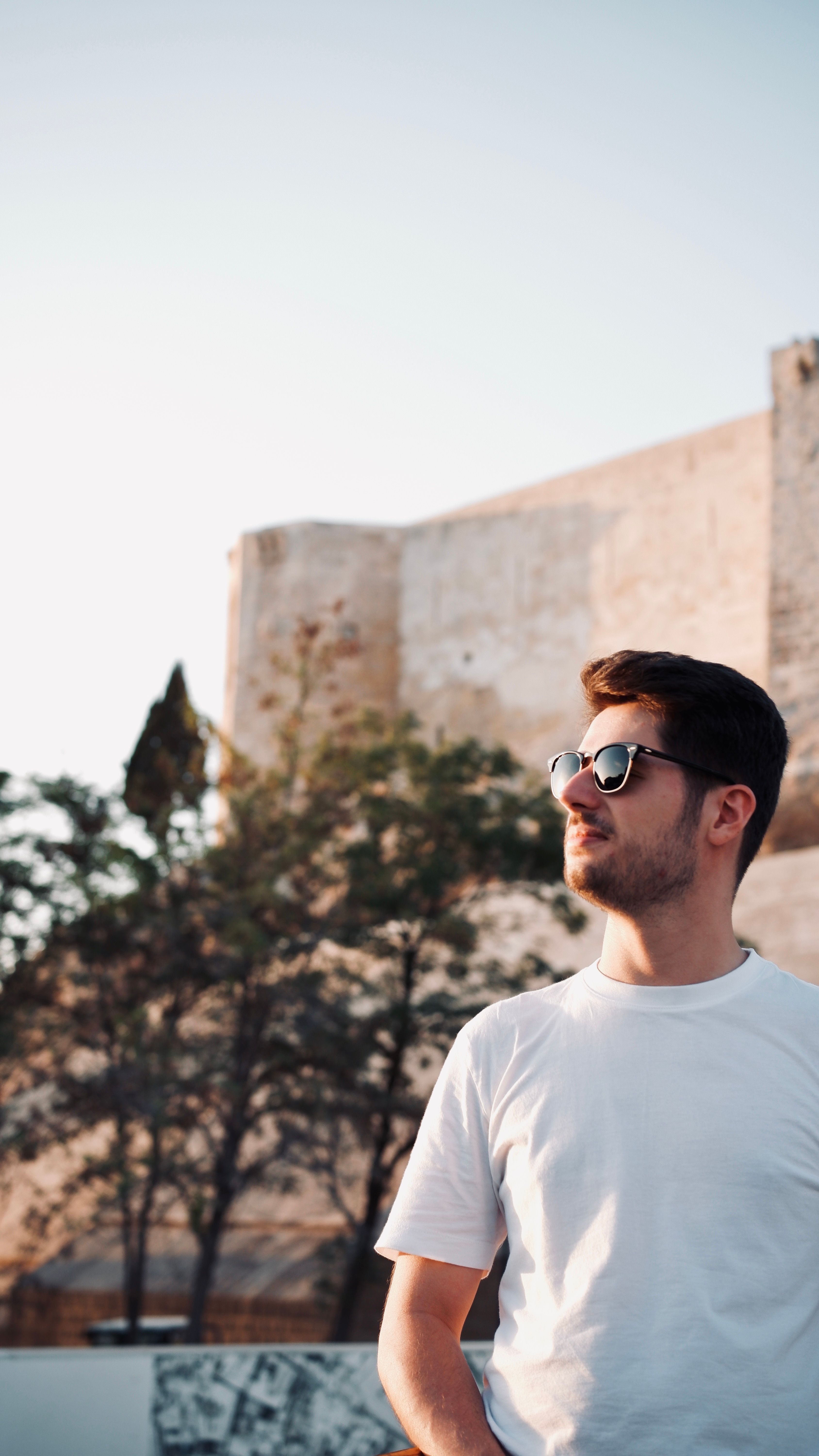 Man in sunglasses outdoors, standing against an old wall, enjoying the sunny day.