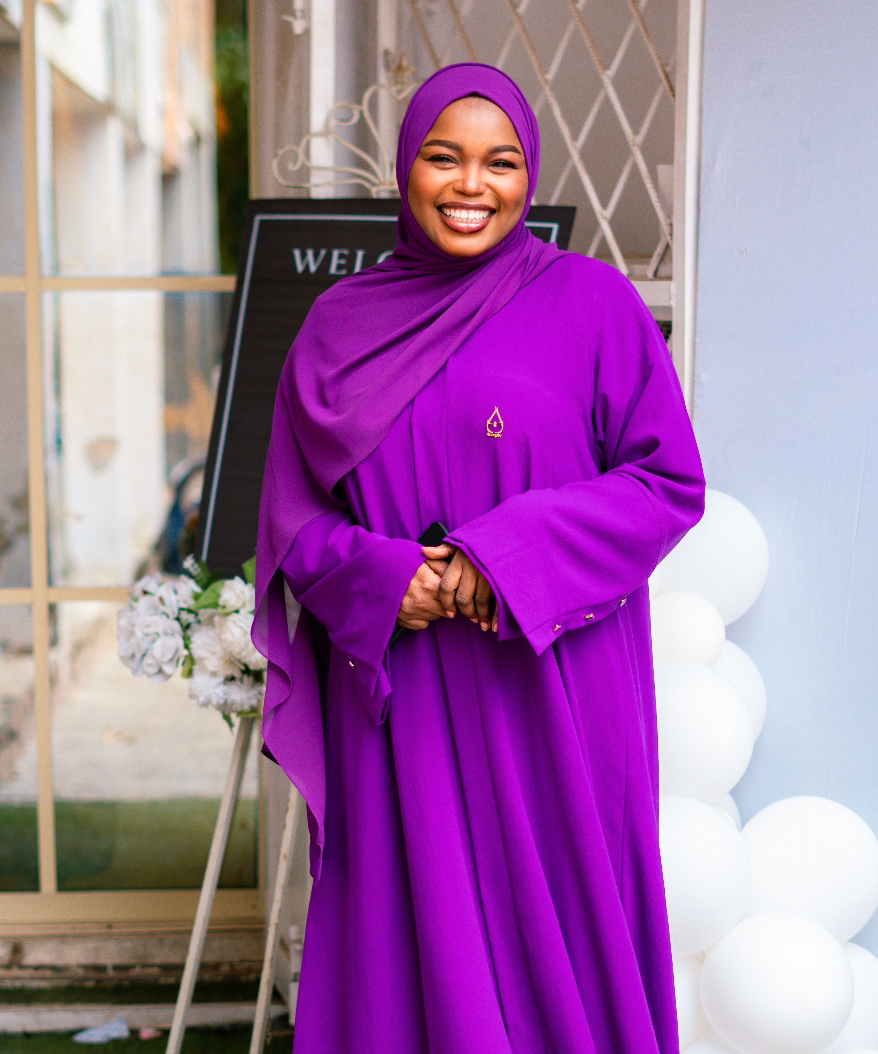 Cheerful woman wearing a purple hijab standing outdoors against a welcoming backdrop.