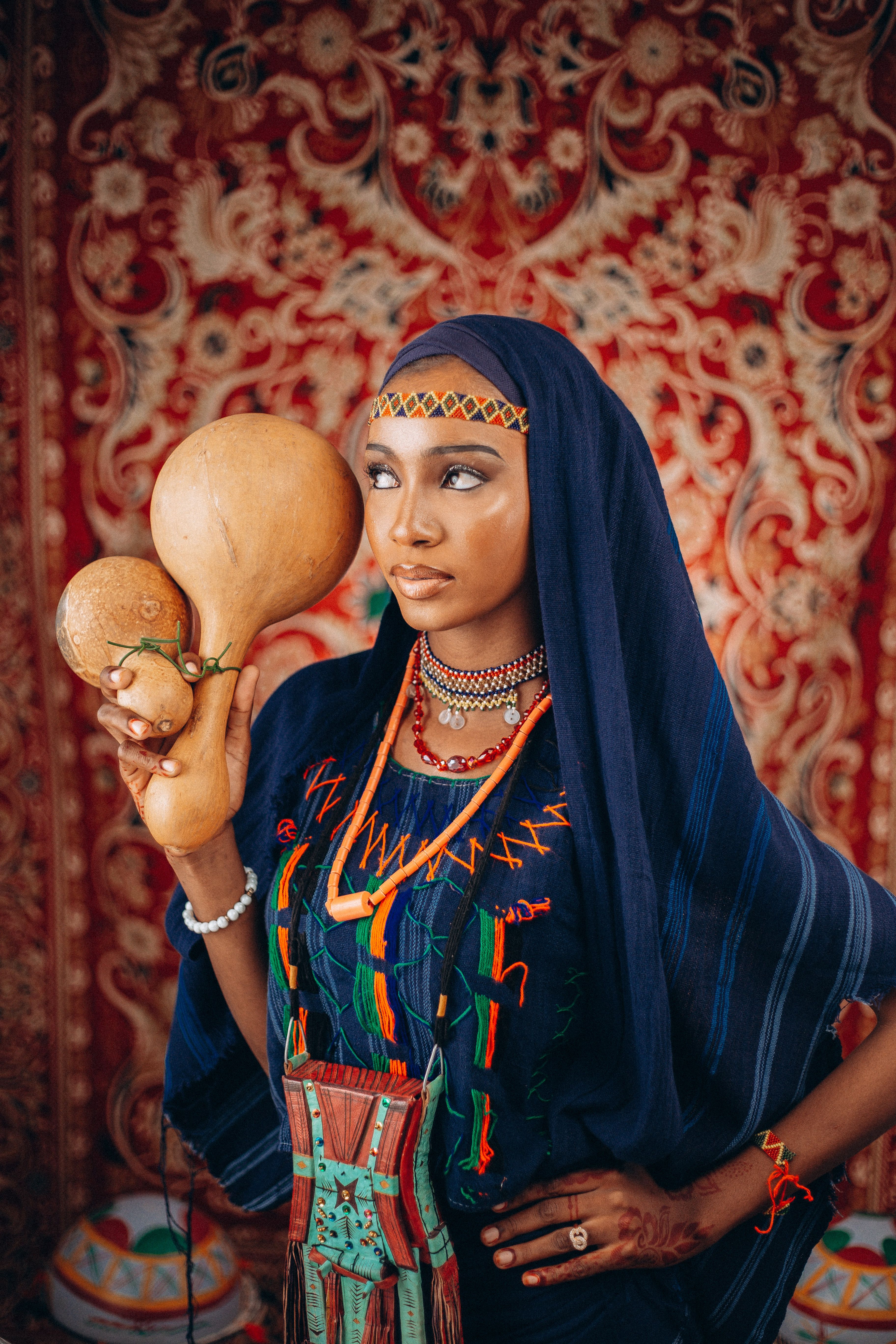 African woman in traditional attire holding calabash against ornate backdrop.
