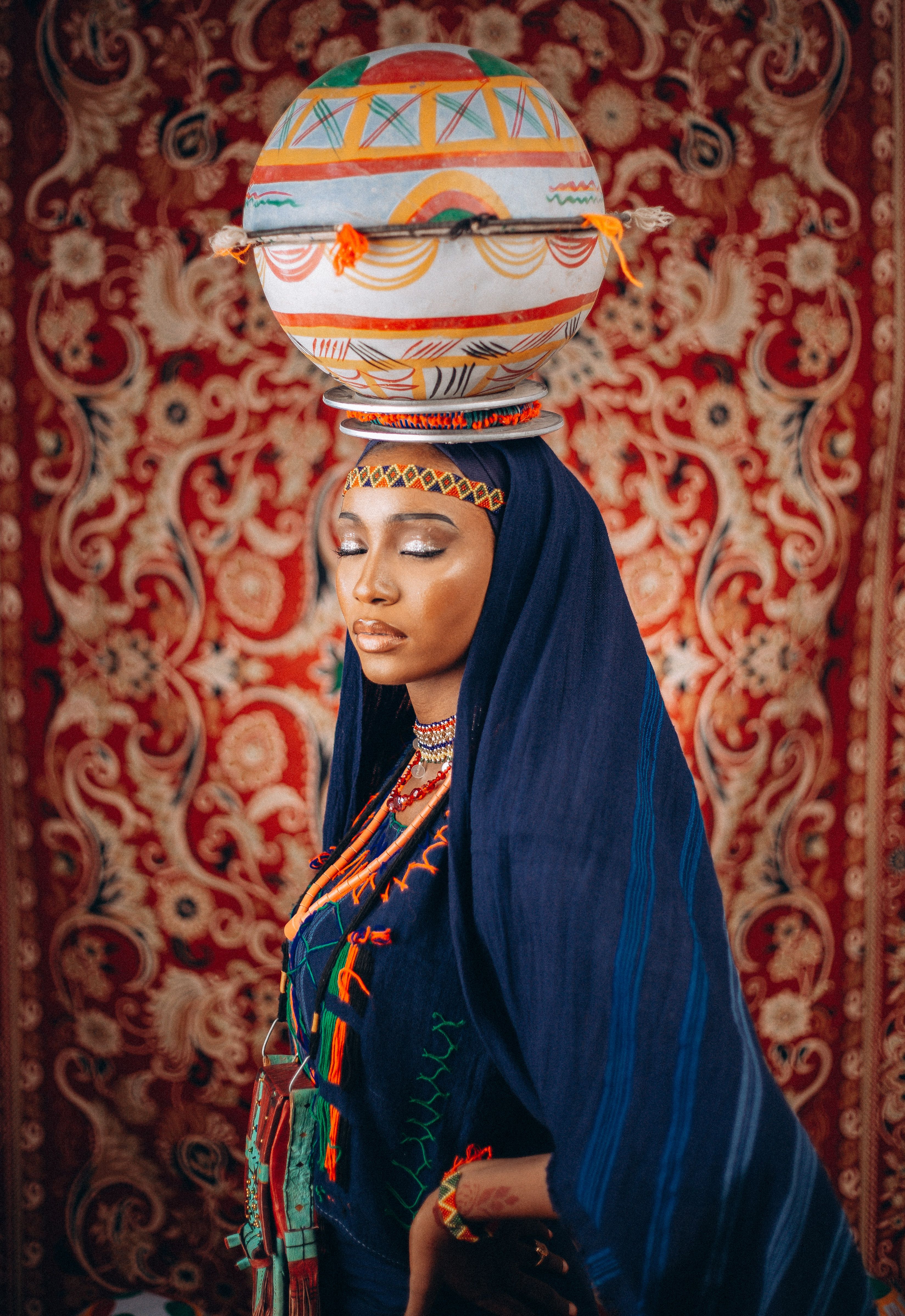 Woman in colorful traditional African attire with a basket headdress against a patterned backdrop.