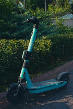 A blue electric scooter stands alone on a brick pathway surrounded by greenery.