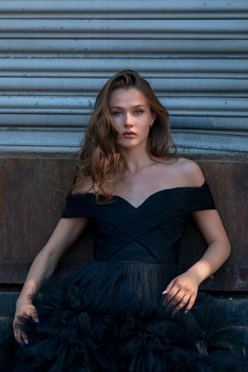 Free Stylish woman in a black off-shoulder dress leans against a textured wall in sunlight. Stock Photo