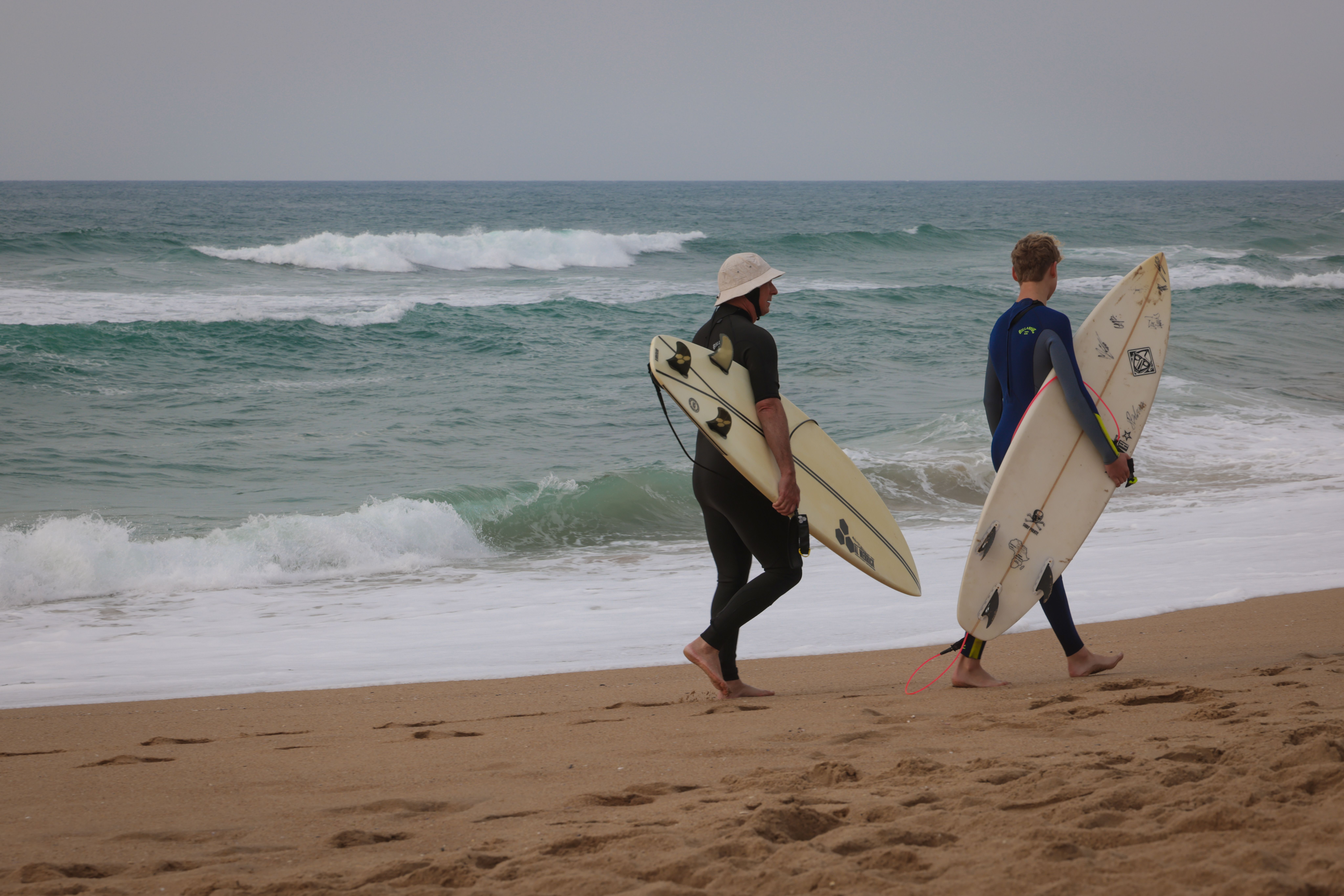 Two surfers carry their boards along a sandy beach with ocean waves in the background.