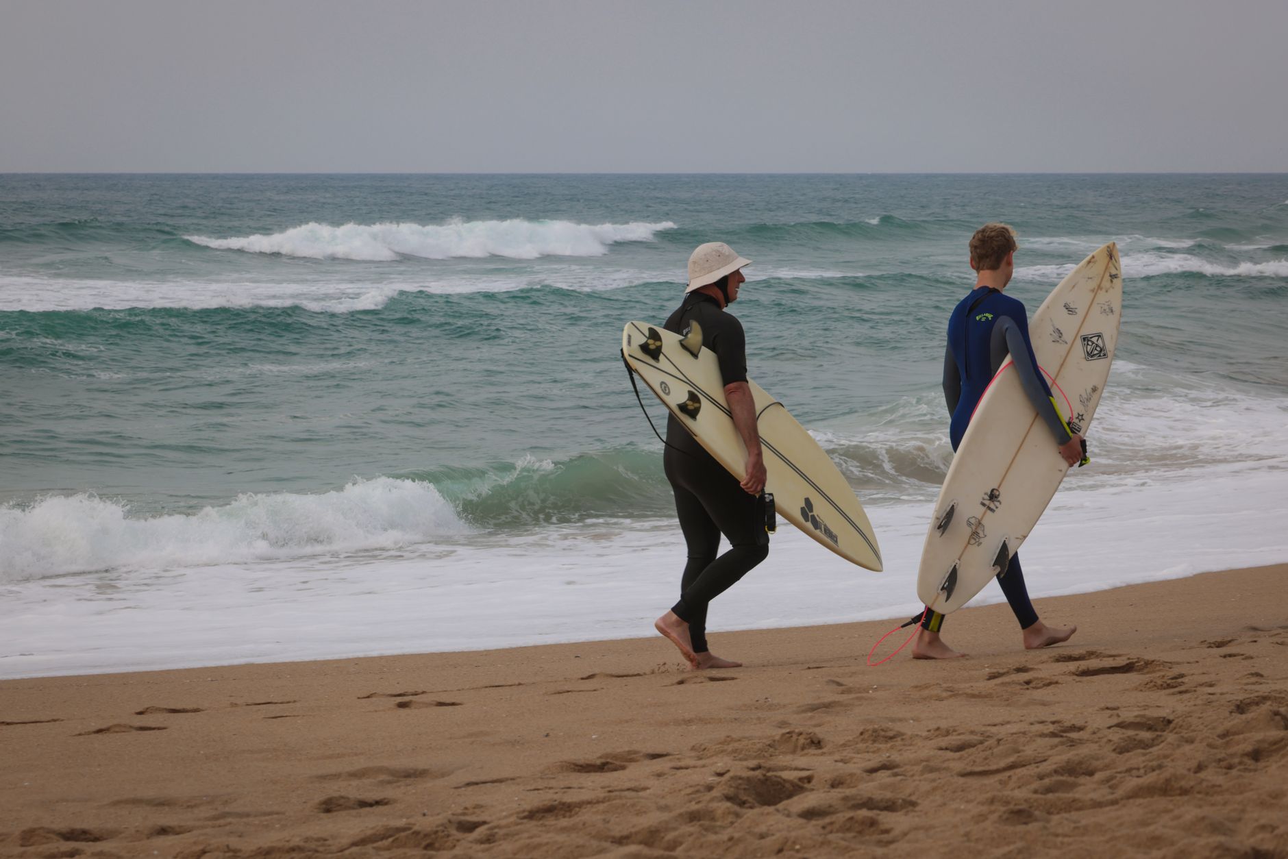 Two surfers carry their boards along a sandy beach with ocean waves in the background.