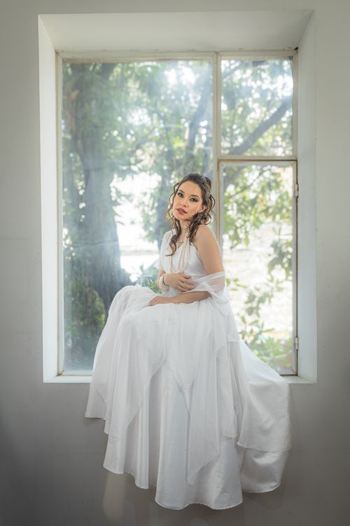 Free A beautiful bride in a flowing white dress poses gracefully by a sunlit window indoors. Stock Photo