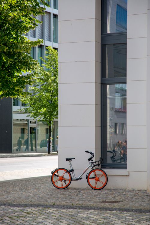 Free Stylish bicycle with orange wheels against a modern city building, hinting at urban life. Stock Photo