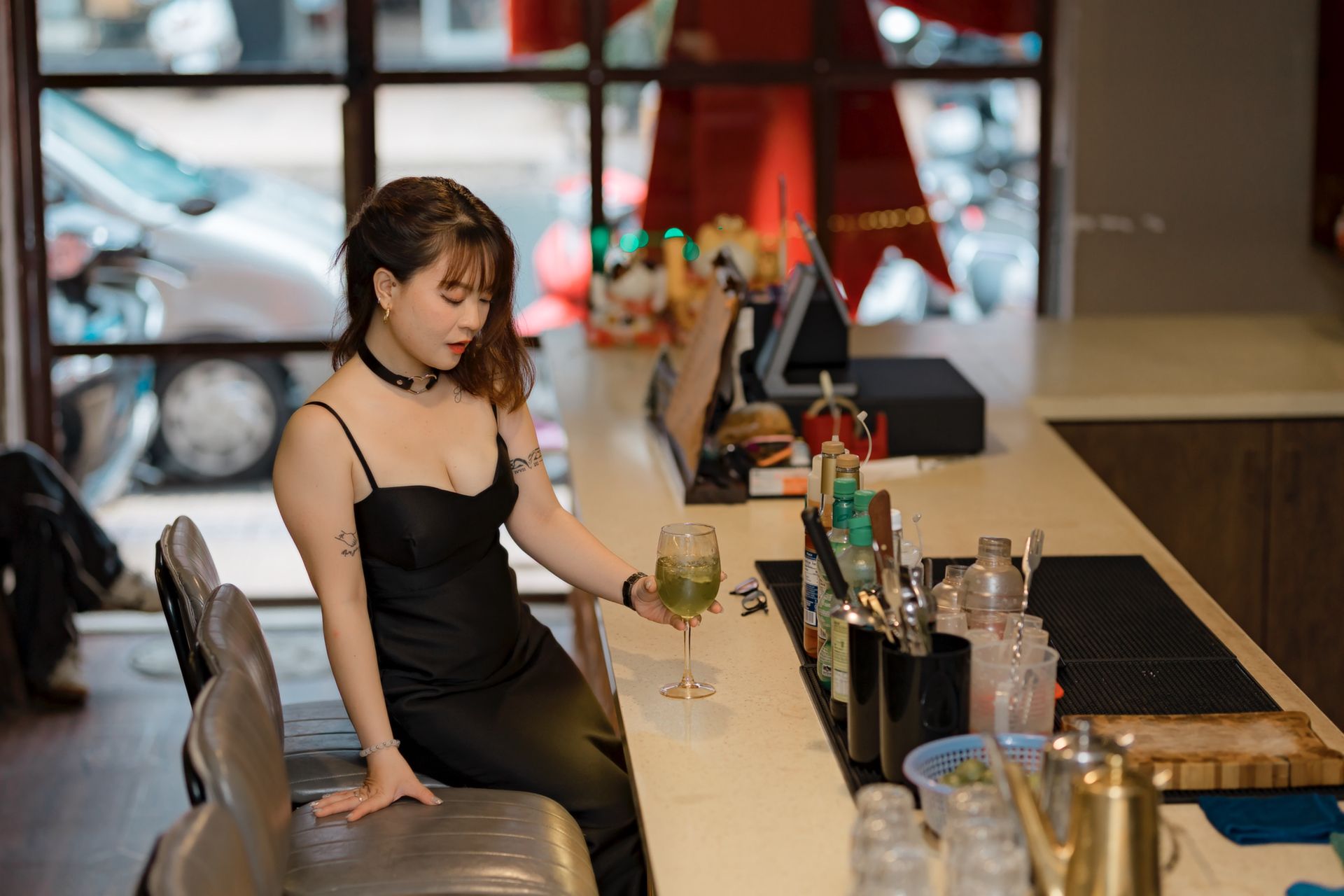 Elegant woman in a black dress enjoying a cocktail at a bar counter indoors.