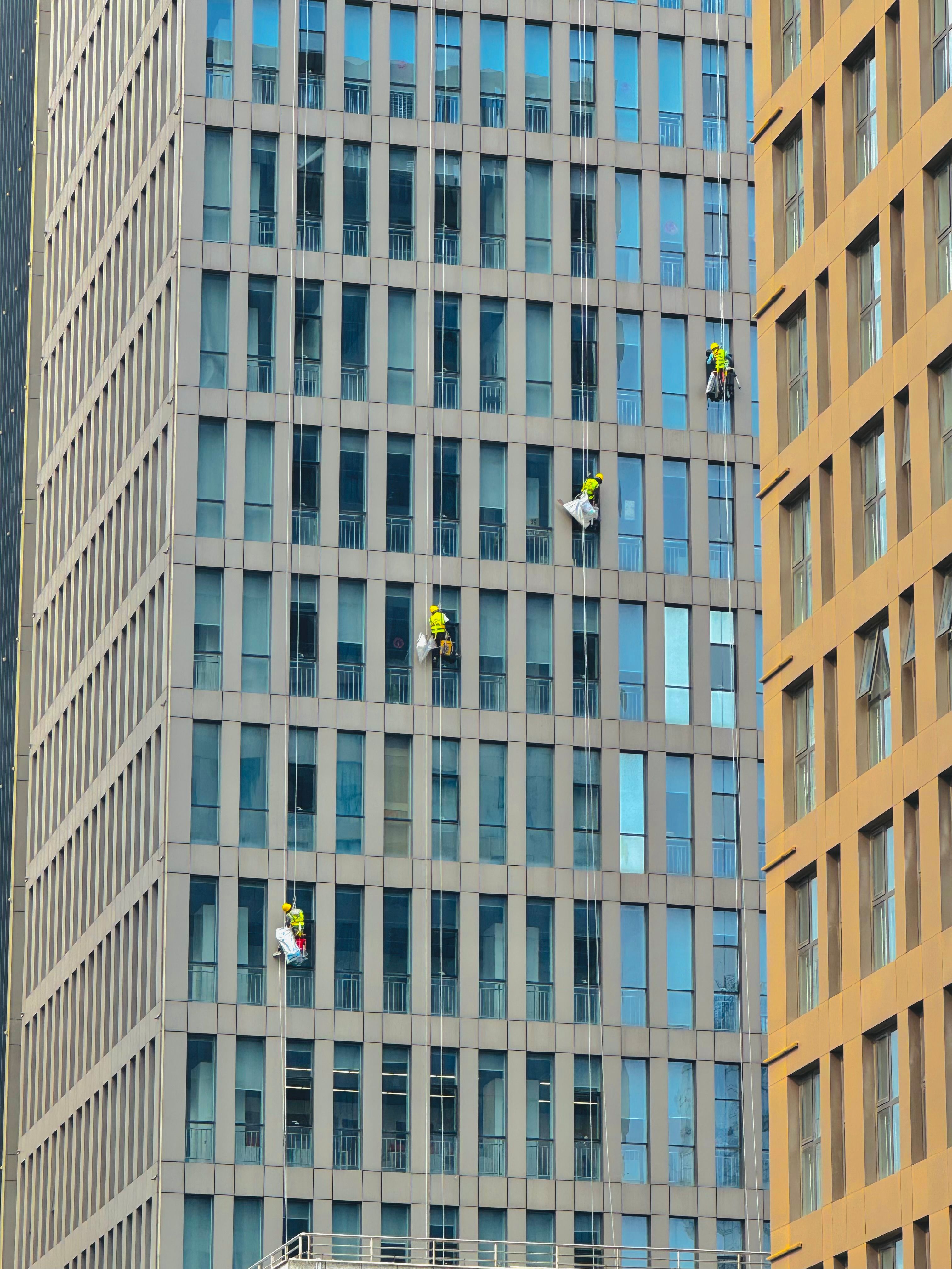 Professional window cleaners suspended on ropes clean a skyscraper facade on a sunny day.