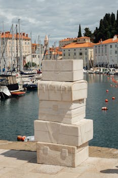 A stone suitcase sculpture by the Piran Harbor, Slovenia, with boats in the background.