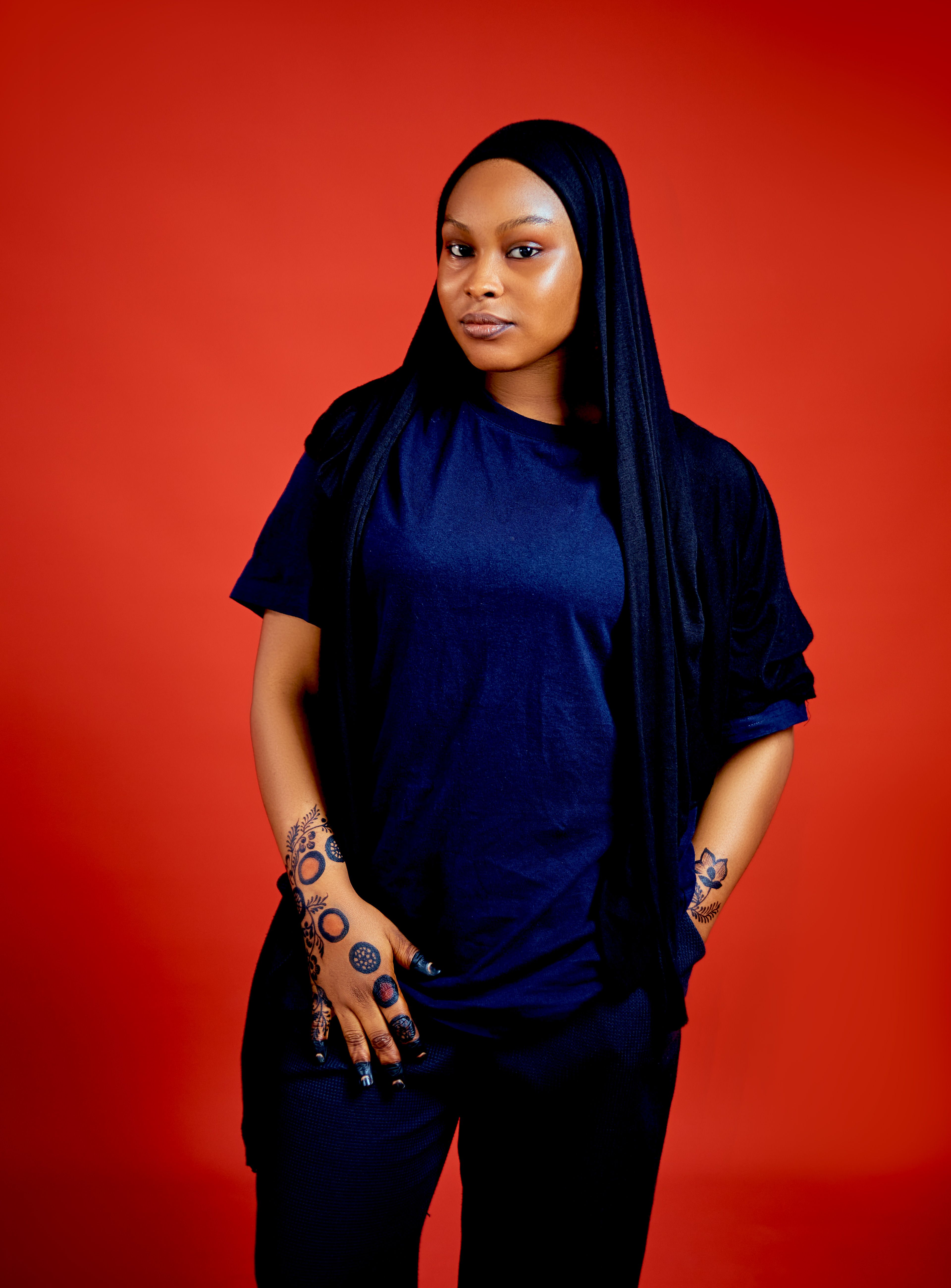 A confident woman with henna tattoos poses in hijab against a striking red background.