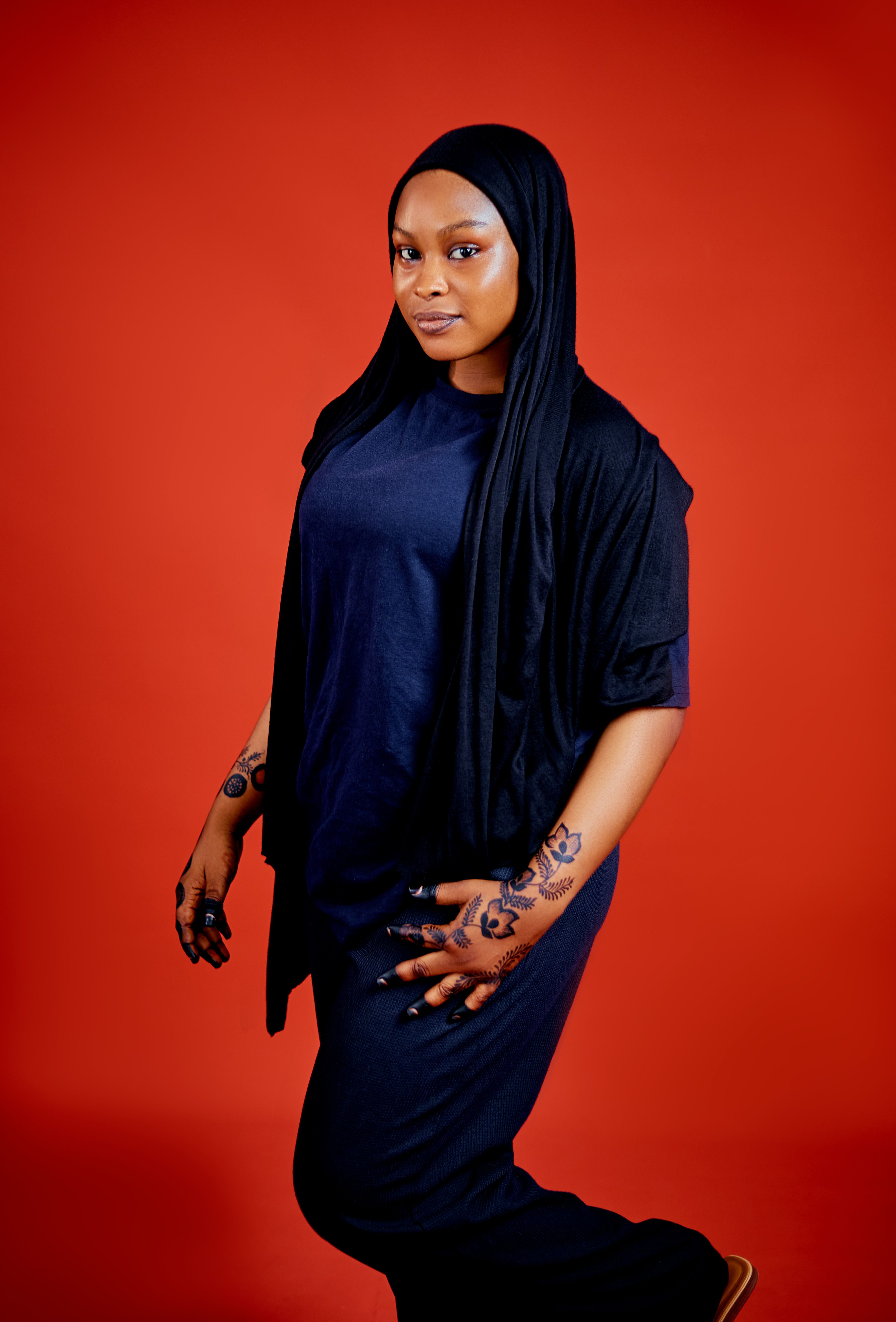 Portrait of a woman in black clothing with henna tattoos, set against a vivid red background.