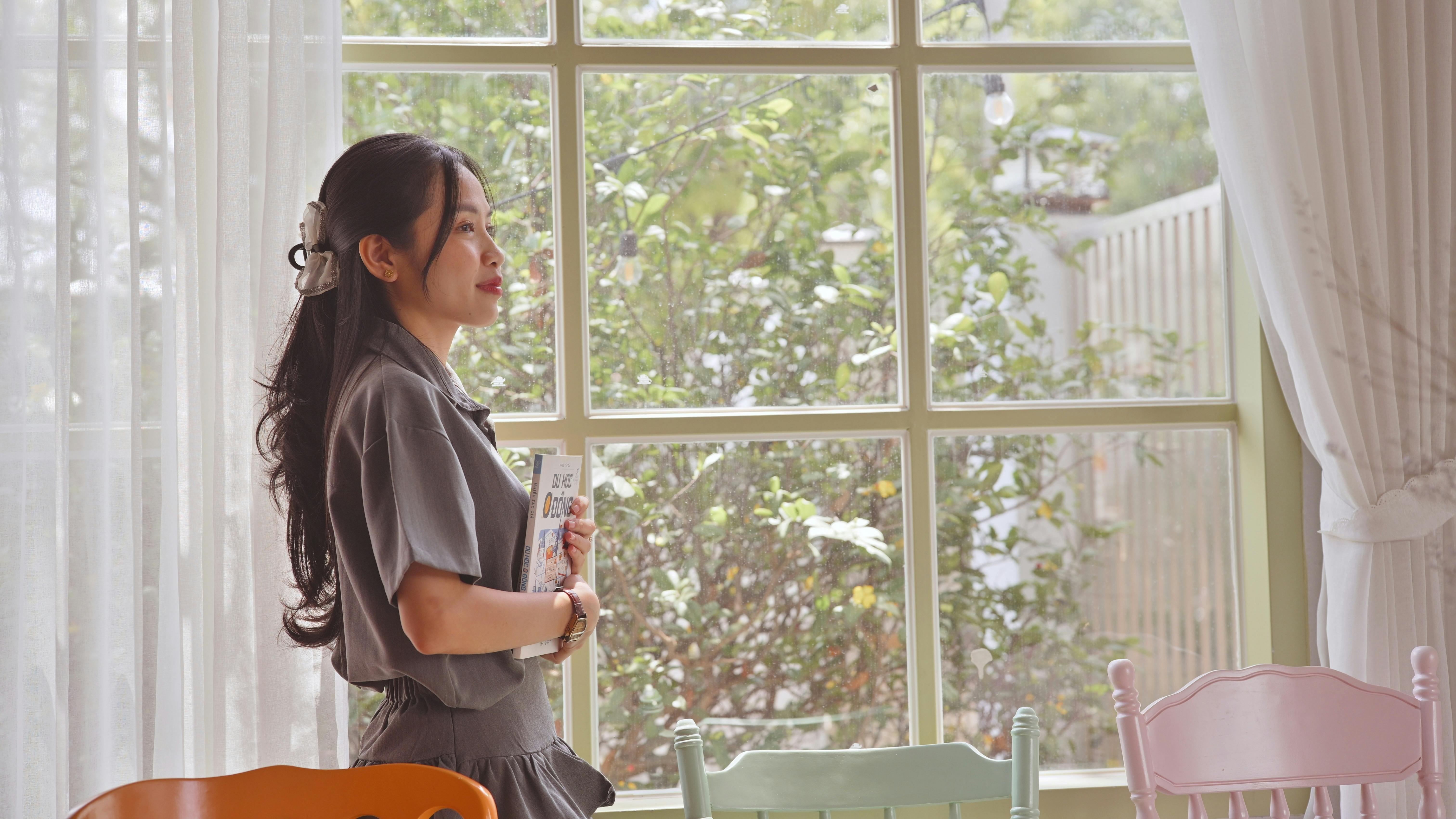 Woman holding a book near a window, enjoying daylight indoors.