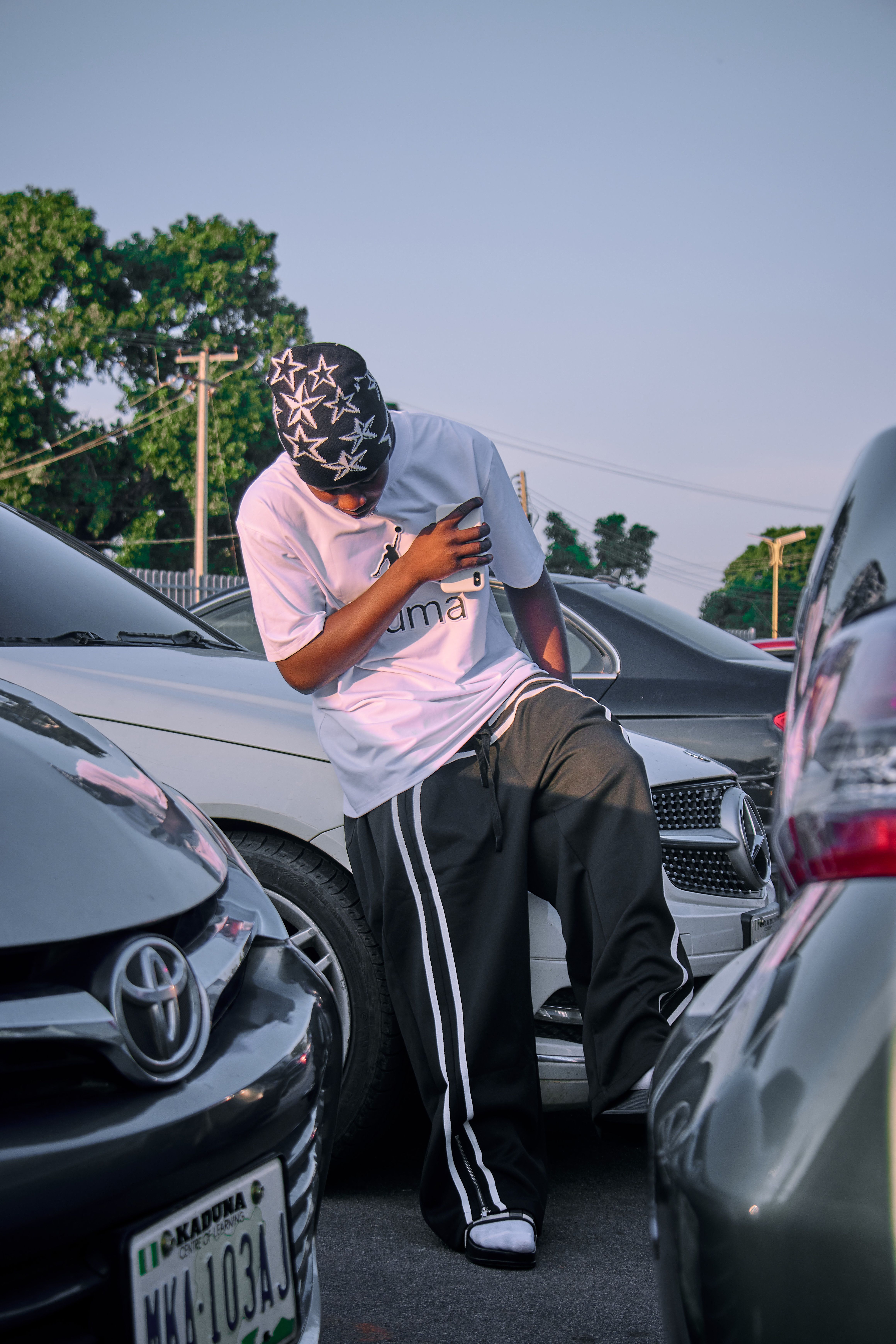 Man standing casually between cars in a parking lot, wearing a stylish street outfit.