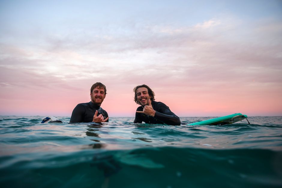 Two surfers in wetsuits enjoy the ocean waves at sunset, giving a shaka sign.