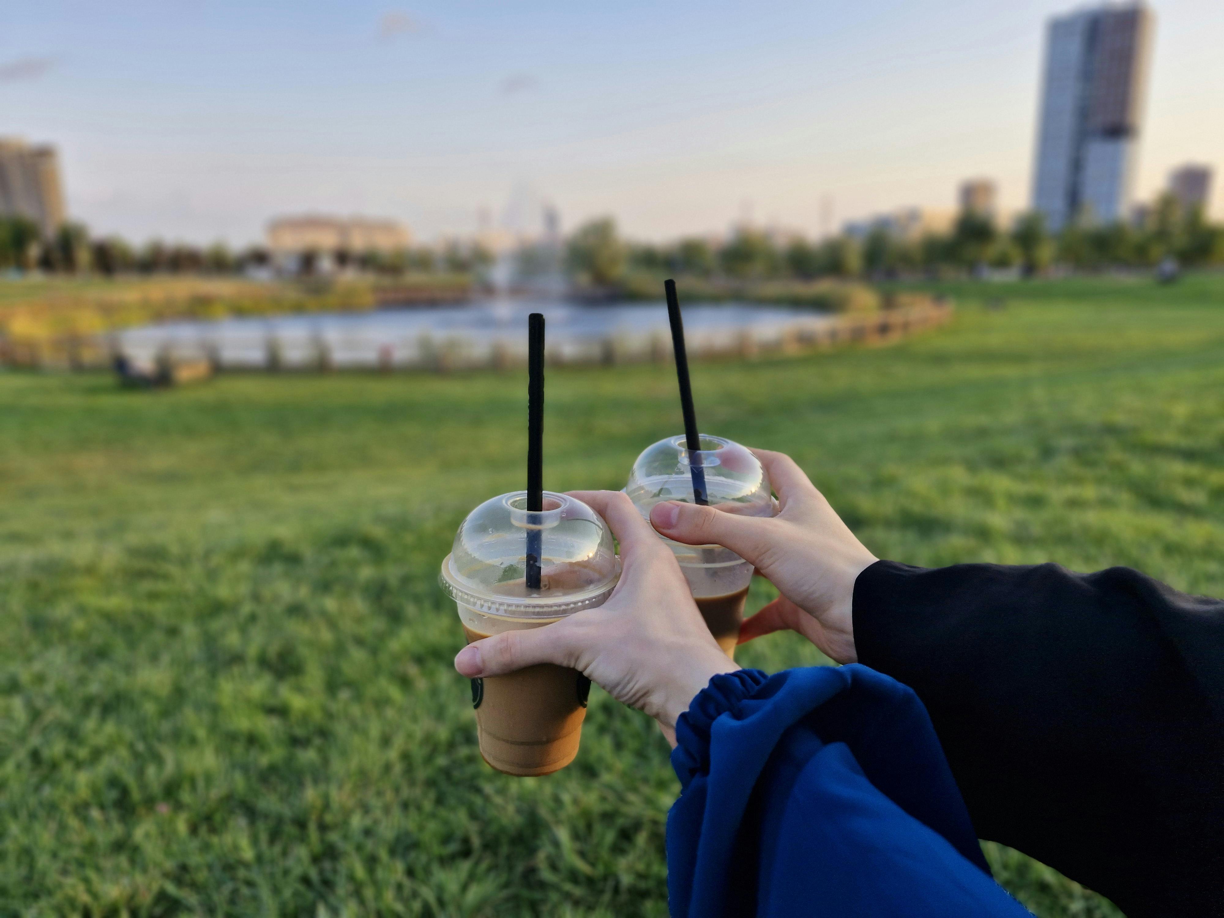 Two people toasting iced coffees in a sunny park, enjoying a lively outdoor setting with cityscape in the background.