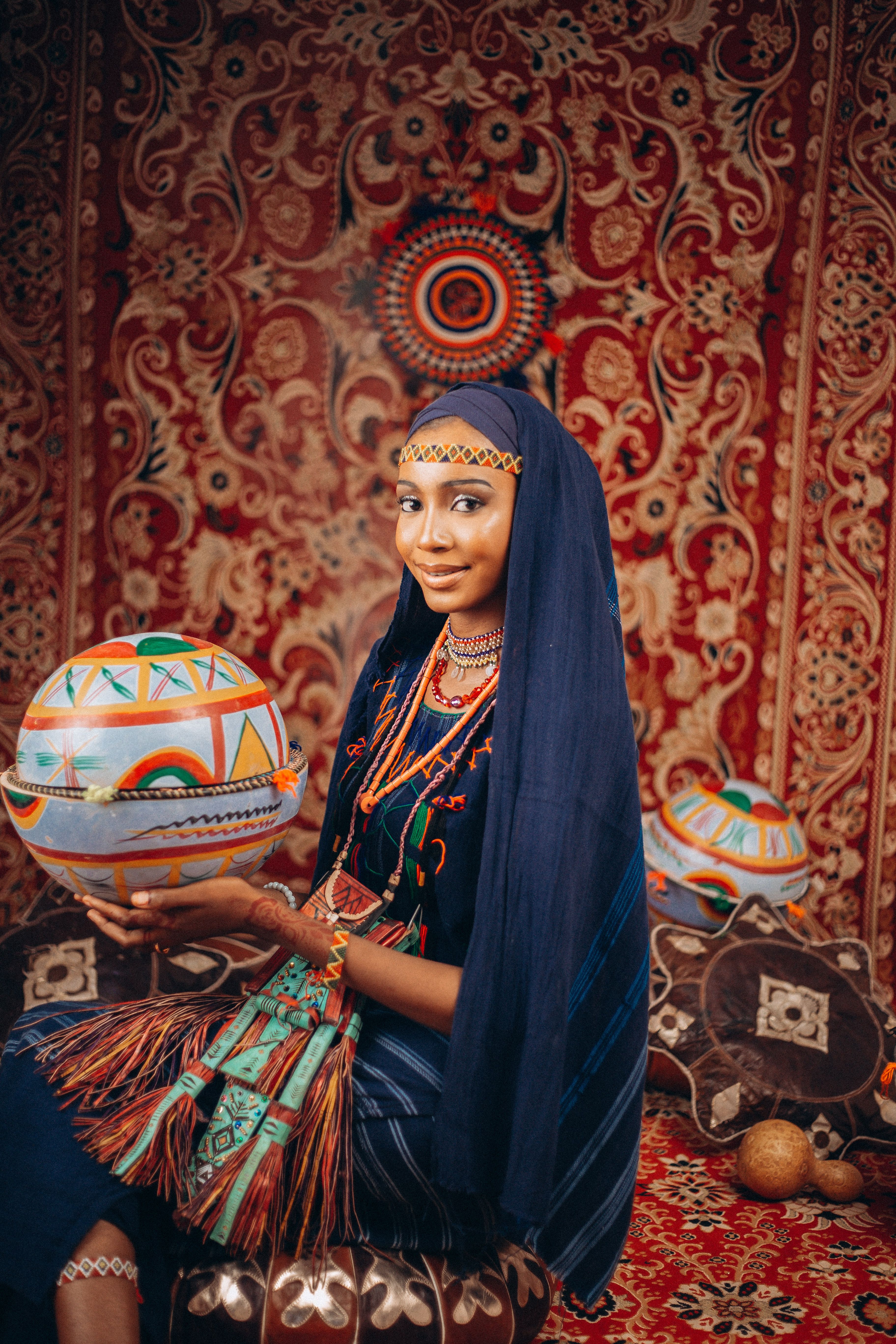 A woman in vibrant traditional attire holding a decorated bowl against rich tapestry.