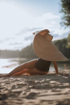 A woman sits on a sandy beach enjoying the sunset, wearing a large straw hat.