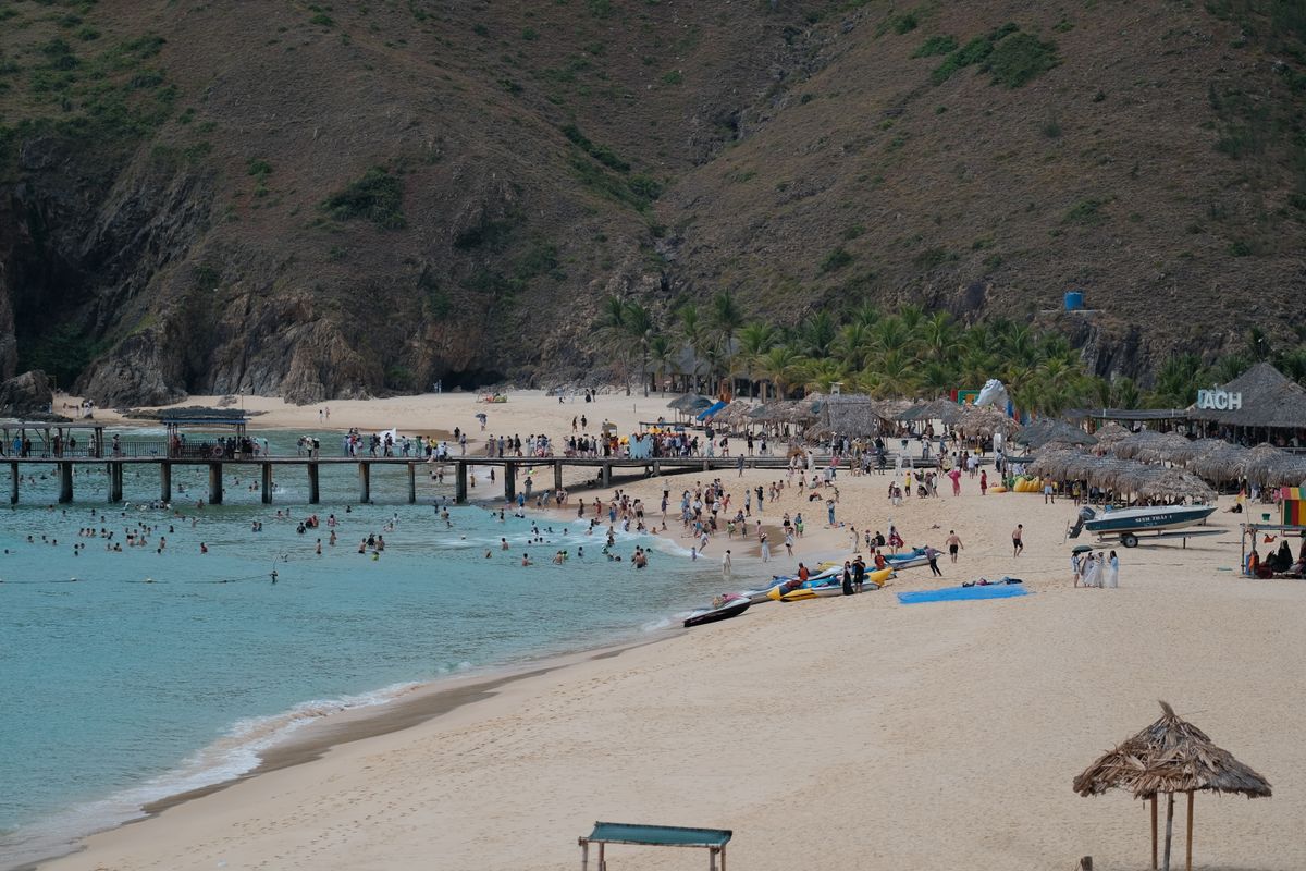 Crowded swimming beach ringed by hills, banana boats lined up along the shore.