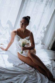 A bride in elegant lingerie poses with a bouquet by a sunlit window, creating a soft, romantic ambiance.