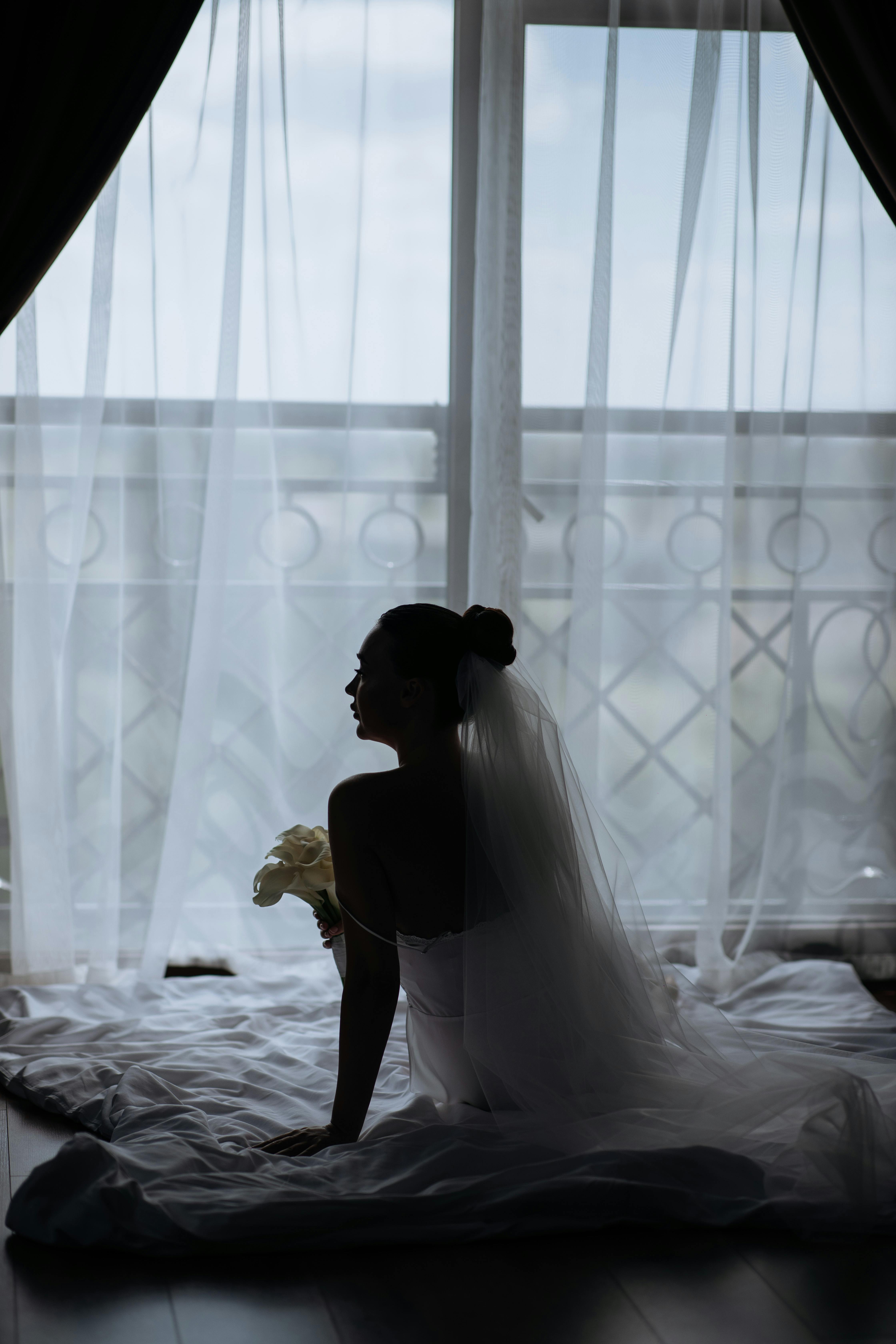 Silhouette of a bride in a veil sitting by a window with soft light filtering through curtains.