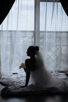 Silhouette of a bride in a veil sitting by a window with soft light filtering through curtains.