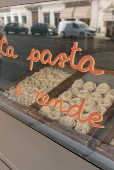 Showcase of Italian pasta varieties behind a shop window, reflecting an urban street scene.